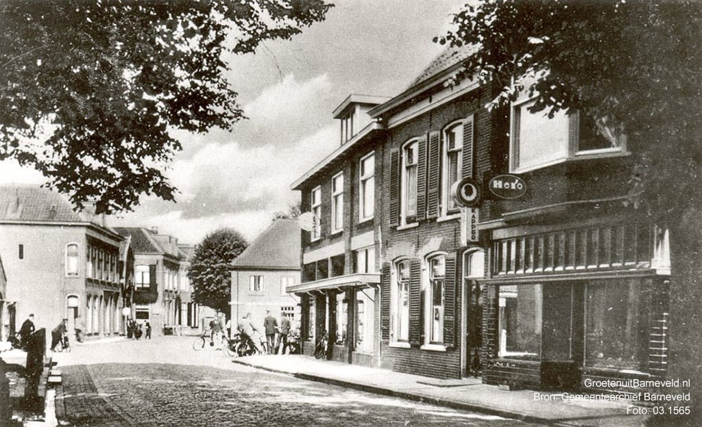 Verleden tussen 1945 en 1955 - De foto is gemaakt in de richting van de Schoutenstraat. Van links naar rechts: de stoep van "Bon Repos", sigarenzaak Schut (voorheen bakkerij Romeijn), voormalige stalhouderij en Hotel De Roskam, Schoutenstraat, smederij W. v.d. Mheen, Gasthuisstraat, modehuis Klomp, de woning van de familie Bos en groentewinkel Hartkamp/kapsalon Leeuwis. - Barneveld