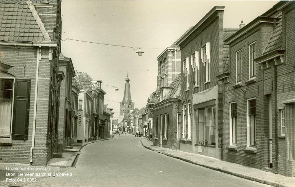 Verleden 1955 - De Langstraat met links het kantoorpand van veevoederfabriek De Heus, iets verder is de zijgevel van schoenenzaak Van der Schans zichtbaar. Rechts de groentenzaak van Kap. In het midden rechts de vakteken- later technische school, nog later werd hier de kledingfabriek Barcon in gevestigd. - Barneveld